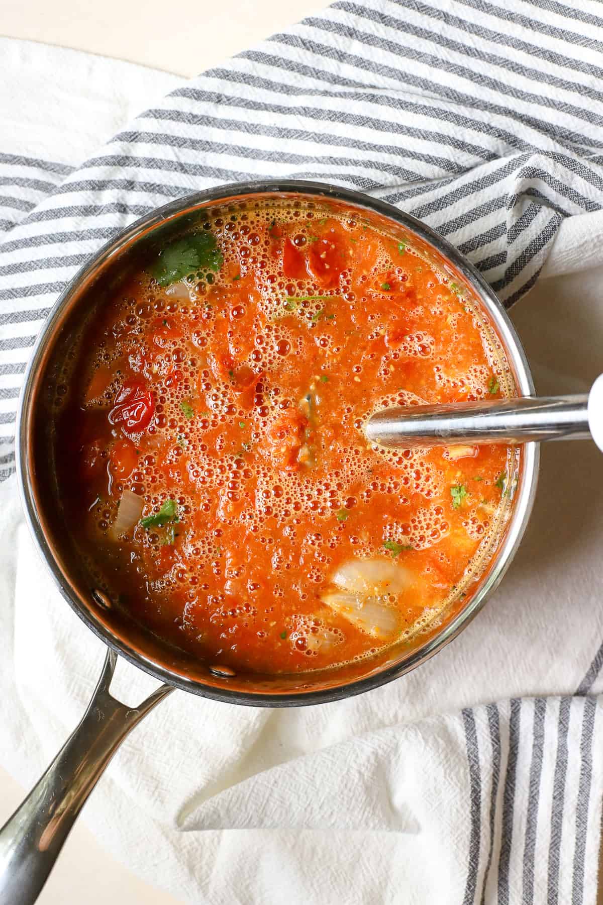 Roasted cherry tomato soup being blended with immersion blender in stainless steel pot.