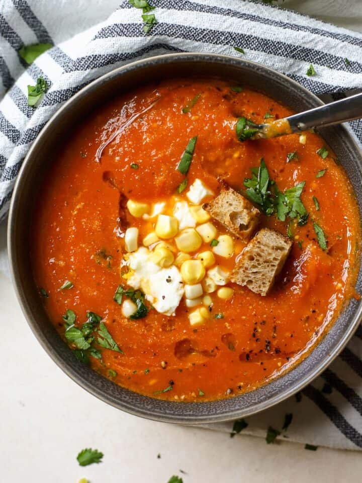 Roasted cherry tomato soup in gray bowl topped with fresh corn, feta, cilantro, pepitas, and croutons.