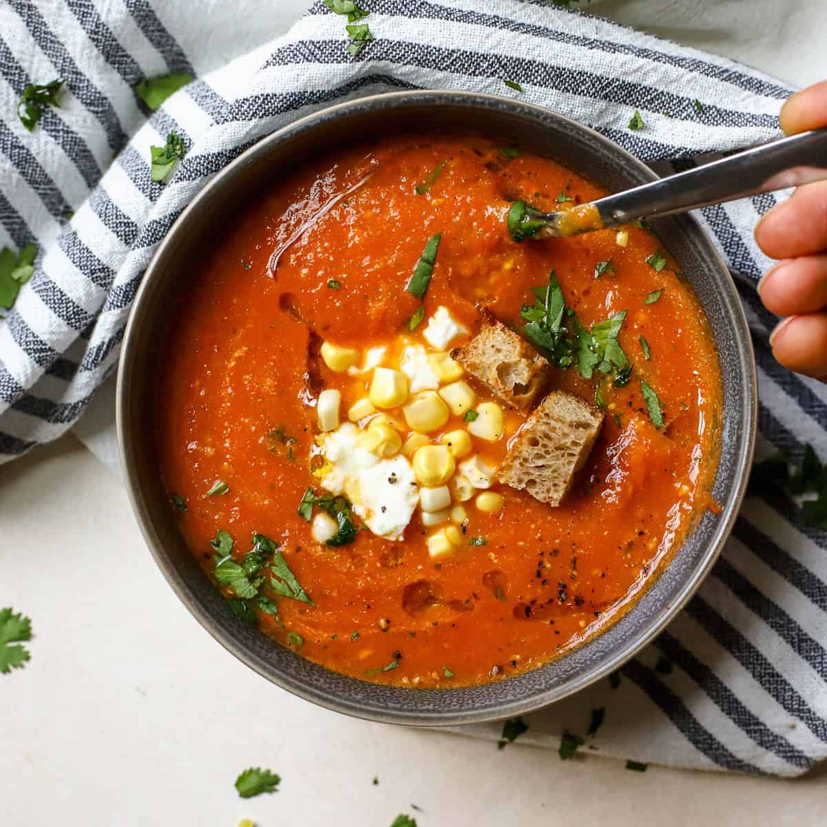 Roasted cherry tomato soup in gray bowl topped with fresh corn, feta, cilantro, pepitas, and croutons.