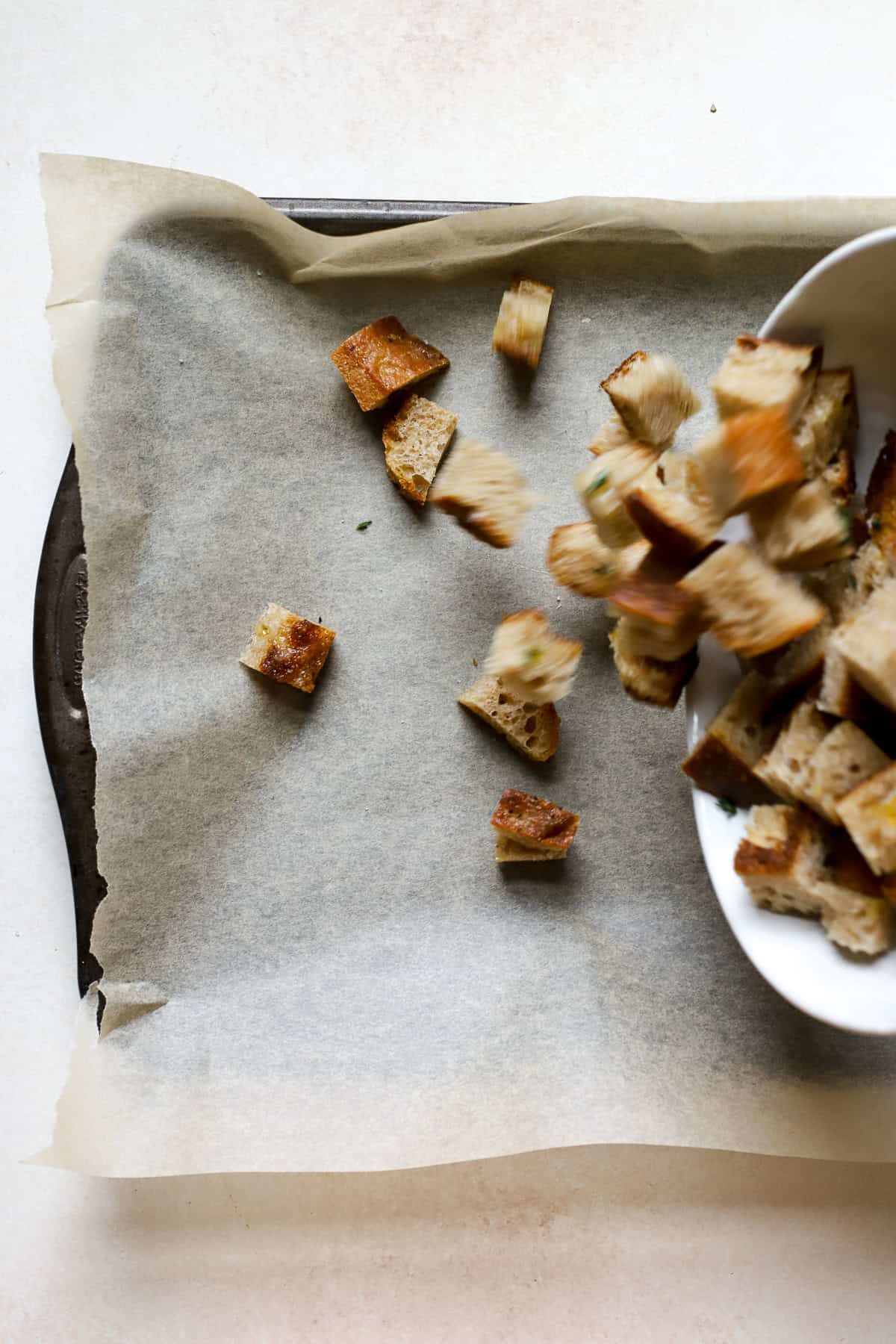 Dumping seasoned sourdough bread cubes onto a parchment-lined sheet pan.