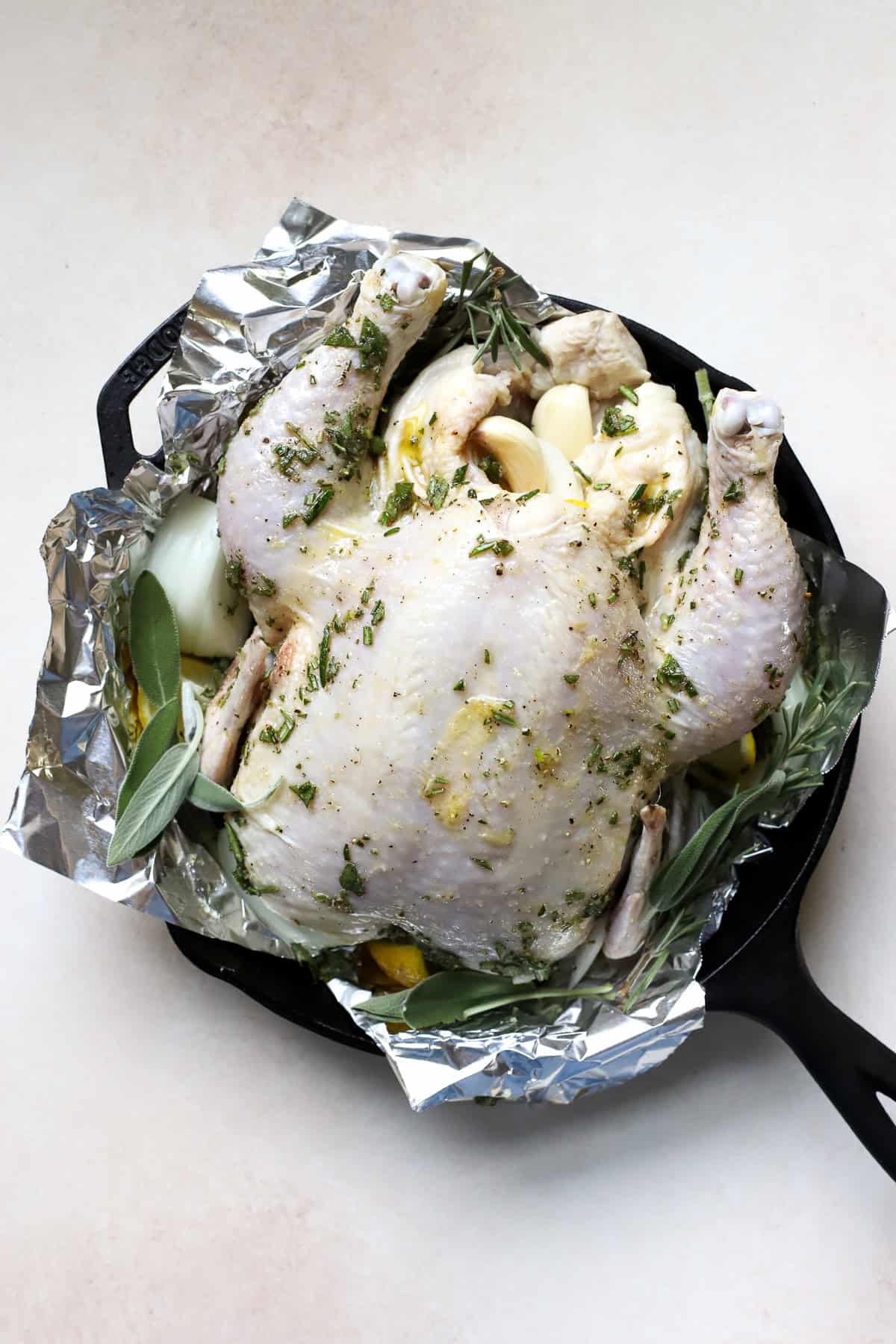 Whole chicken in skillet prior to roasting, seasoned with olive and fresh herbs.