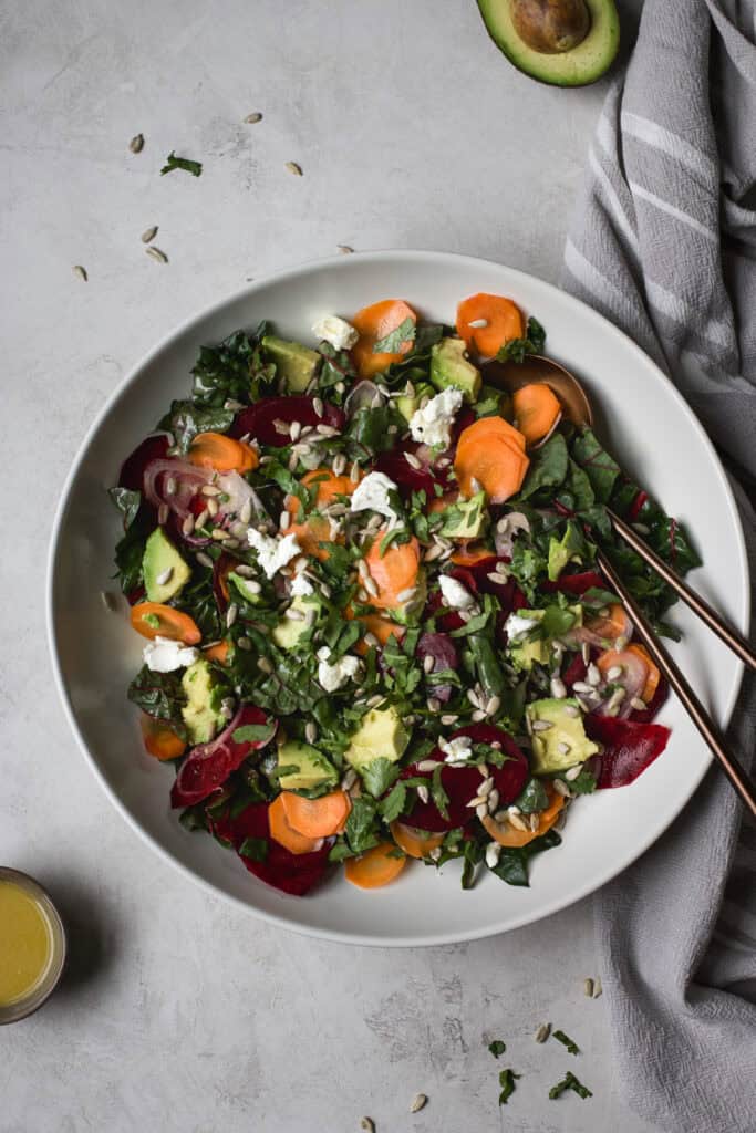 Greens salad with beets and carrots in large white serving bowl with serving spoons.