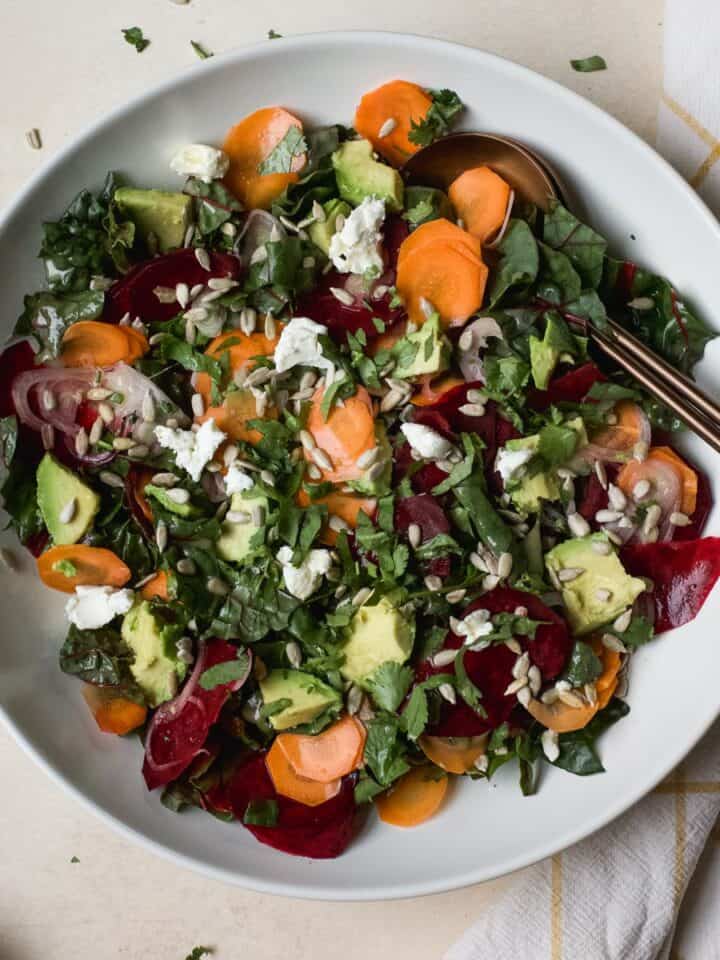 Greens salad with beets and carrots in large white serving bowl with serving spoons.