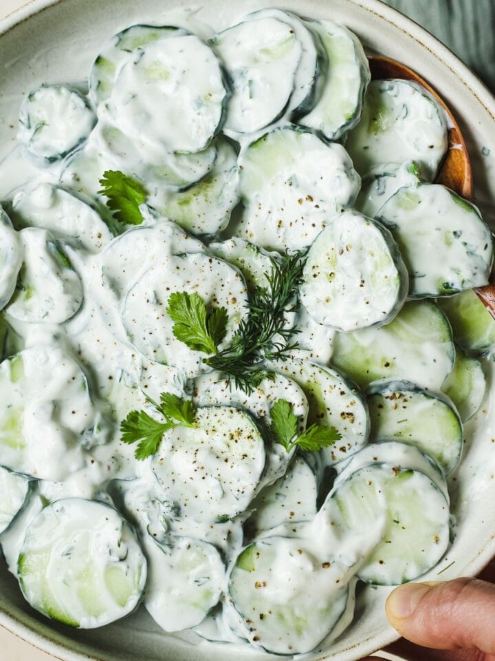 Hand holding creamy cucumber salad in serving bowl.