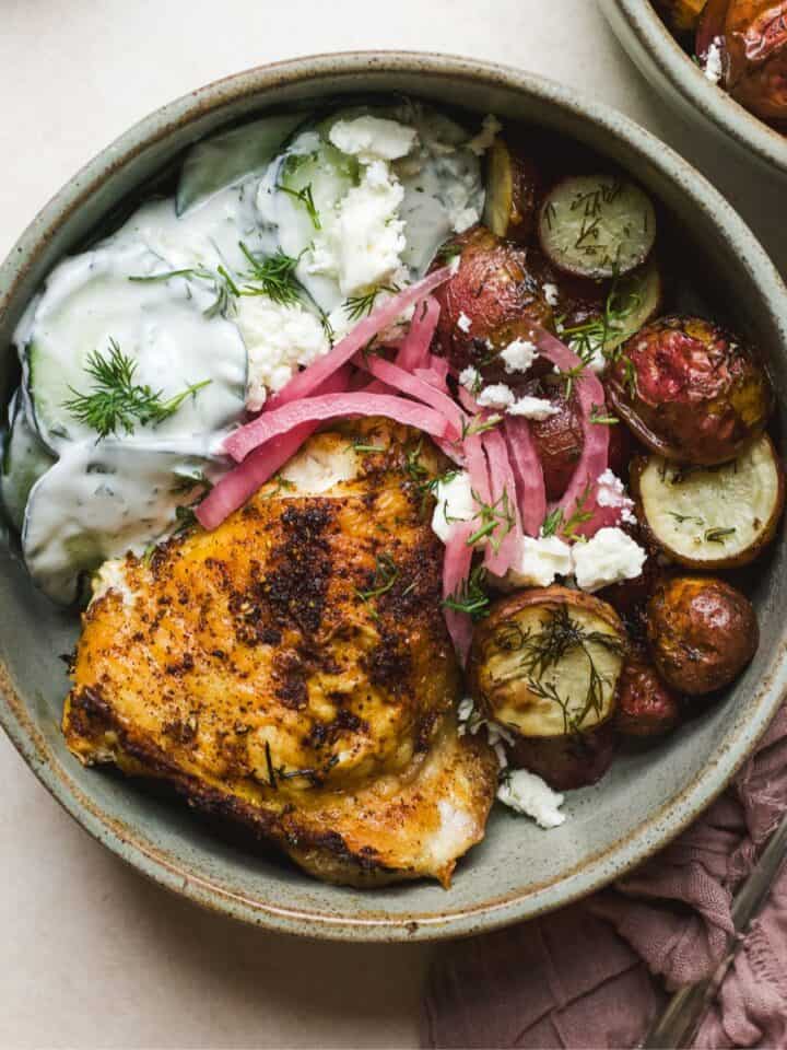 Berbere chicken with dill roasted potatoes in a bowl with creamy cucumber salad and pickled red onions.