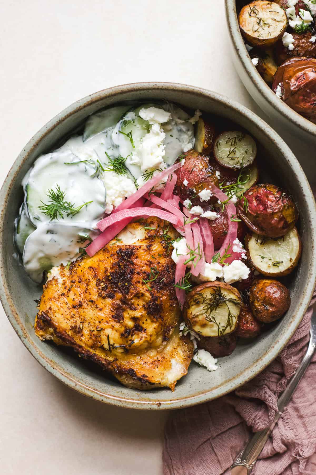 Berbere chicken with dill roasted potatoes in a bowl with creamy cucumber salad and pickled red onions.