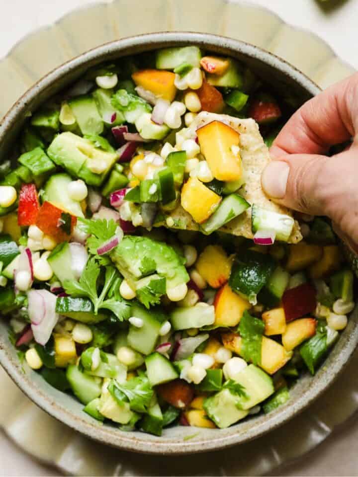 Hand dipping a tortilla chip into fresh peach salsa with corn and avocado.