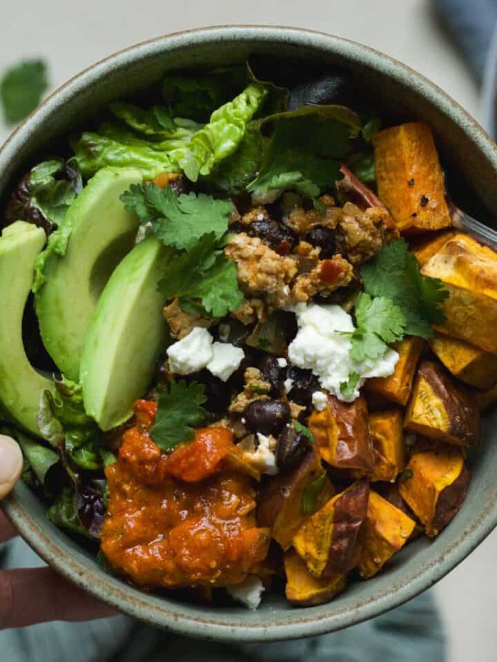 Hand holding a ground chicken taco sweet potato bowl topped with feta, avocado, and cilantro.