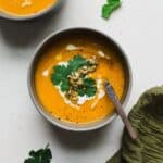 Bowl of spiced carrot and sweet potato soup with a creamy swirl on top, with cilantro and seeds.