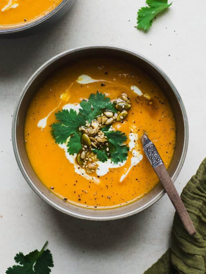 Bowl of spiced carrot and sweet potato soup with a creamy swirl on top, with cilantro and seeds.