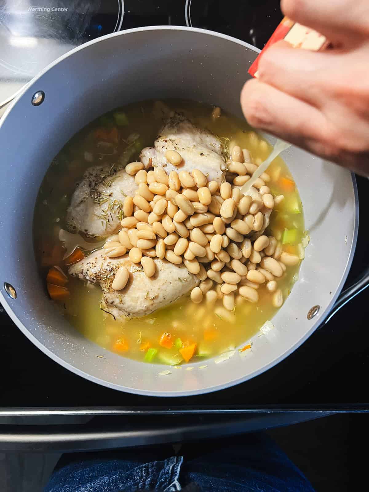 Adding white beans, broth, rosemary, and chicken to soup pot.
