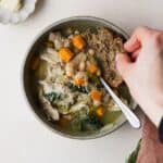 Rosemary chicken soup with kale and white beans, in a bowl and hand dunking fresh sourdough bread into the soup.