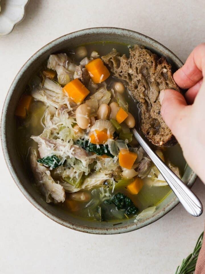 Rosemary chicken soup with kale and white beans, in a bowl and hand dunking fresh sourdough bread into the soup.