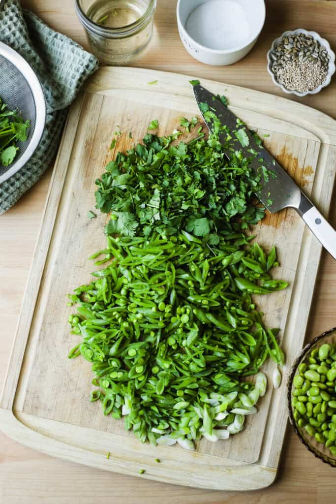 Sliced snap peas and green onions with chopped cilantro on a cutting board with a chef's knife.