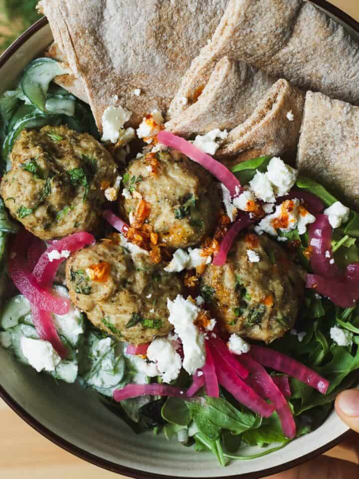Herby chicken meatball bowls with pita wedges, greens, creamy cucumber salad, feta, and pickled red onions.