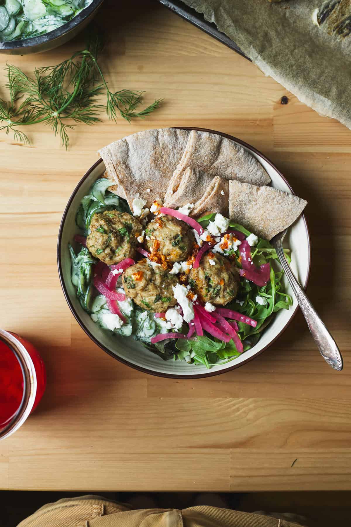 Herby chicken meatball bowls with pita wedges, greens, creamy cucumber salad, feta, and pickled red onions.