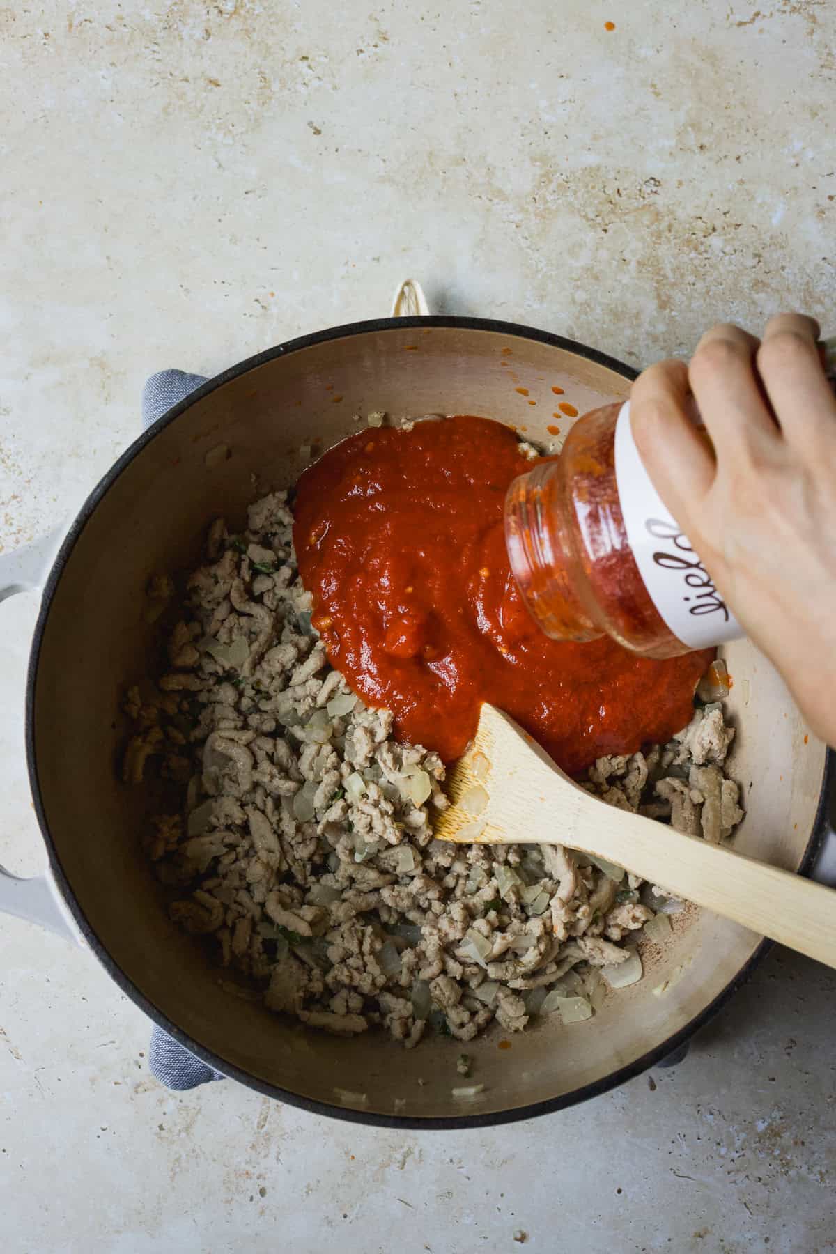 Pouring marinara sauce into a pot with ground meat and aromatics.